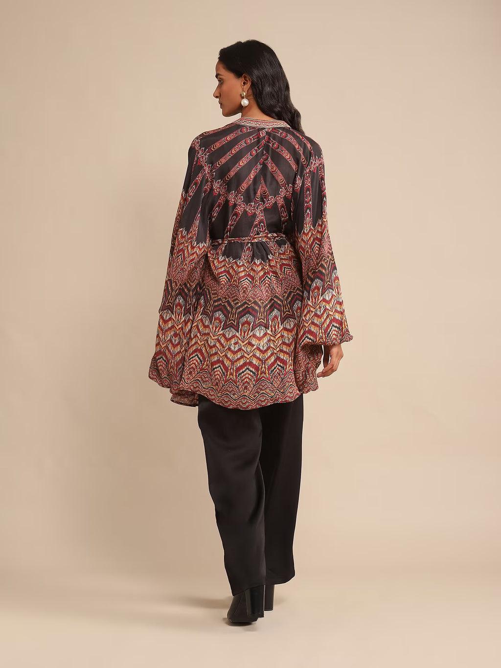 Woman in a printed kaftan-style tunic with flared sleeves, paired with black pants and heels, posing against a beige background.