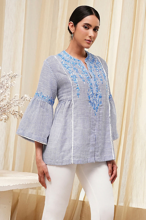 Woman wearing a blue and white striped blouse with embroidery, standing in front of beige curtains.