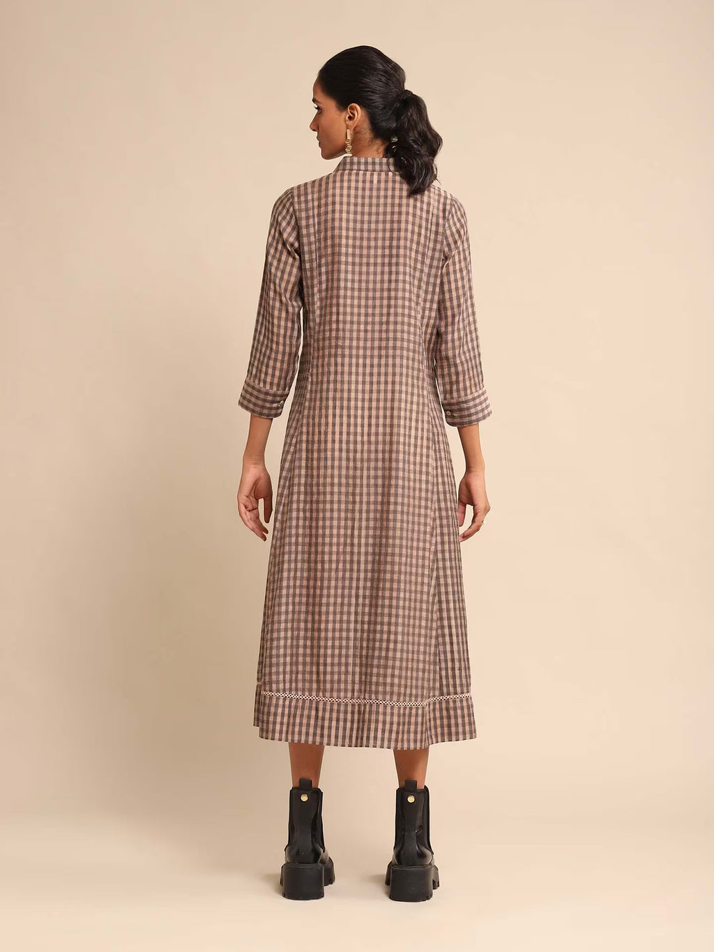 Woman in a brown and beige checked A-line kurta dress with a shirt collar and three-quarter sleeves, paired with black boots, standing against a beige background