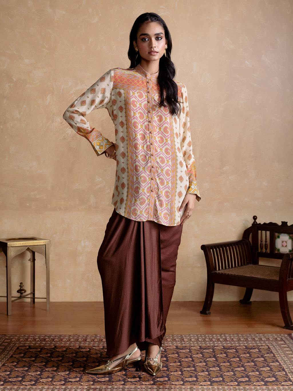 Woman wearing a patterned top and brown dhoti standing in a room with a beige wall and wooden furniture.