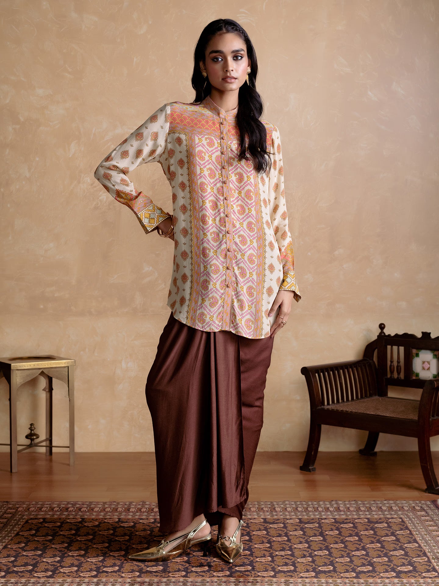 Woman wearing a patterned top and brown dhoti standing in a room with a beige wall and wooden furniture.