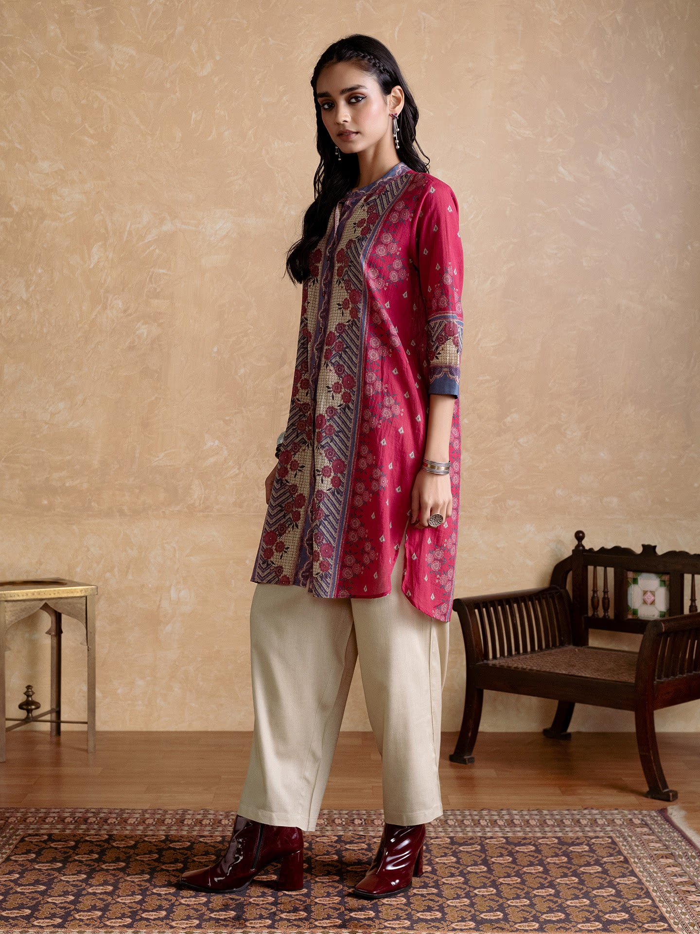 Woman wearing a red and patterned kurta with white pants in a room with wooden furniture and a beige wall.
