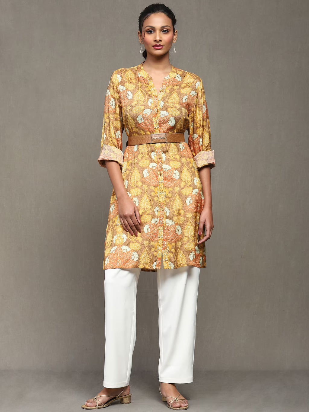Woman wearing a mustard floral printed kurta with a tan belt, paired with white straight pants and heels, standing against a plain grey background.