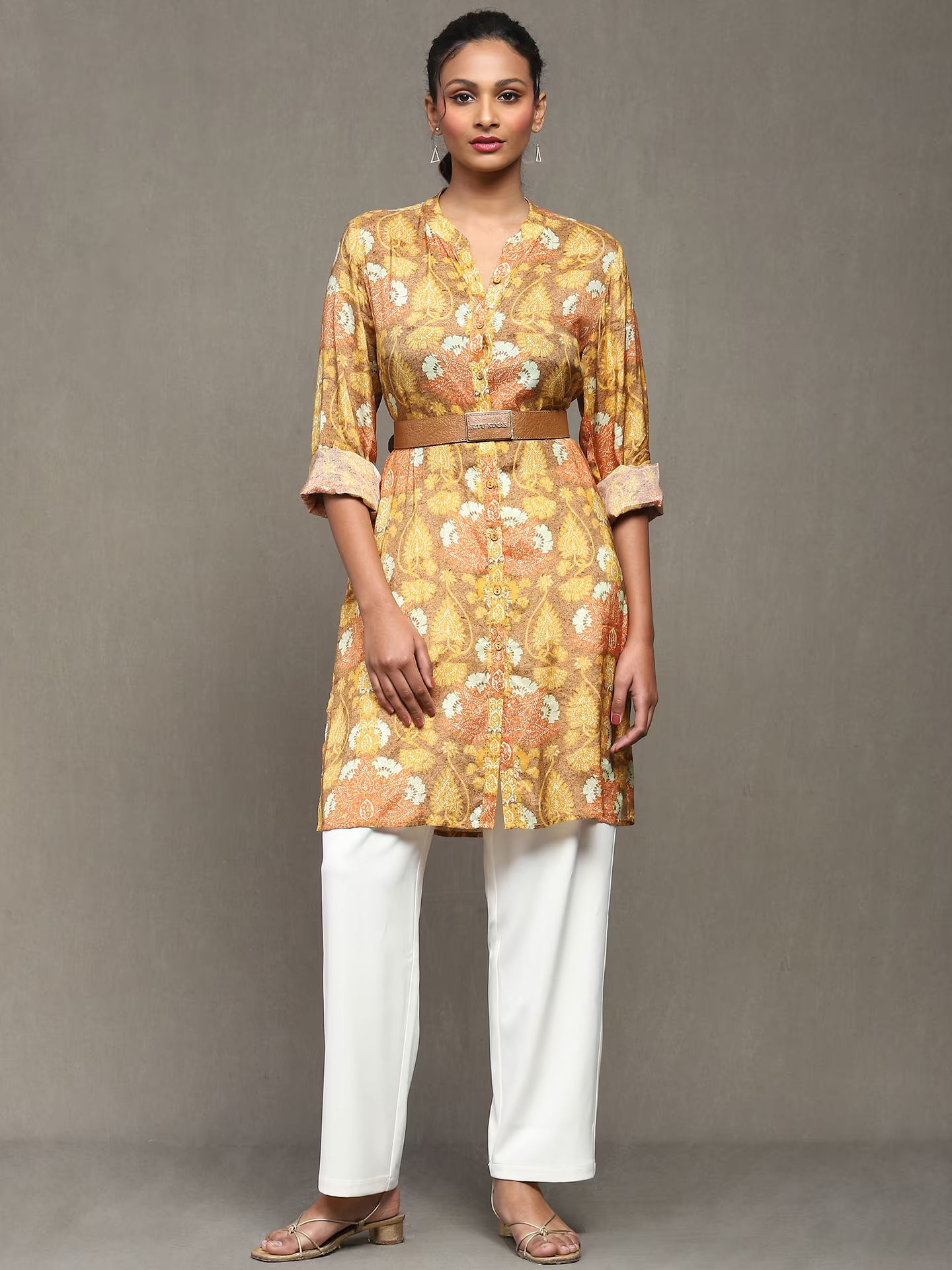 Woman wearing a mustard floral printed kurta with a tan belt, paired with white straight pants and heels, standing against a plain grey background.
