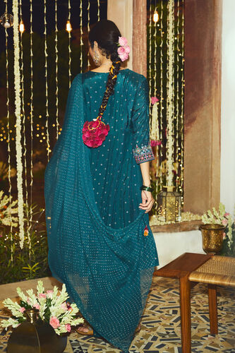 Woman in a teal traditional outfit with a pink floral accessory, standing in a decorated room.