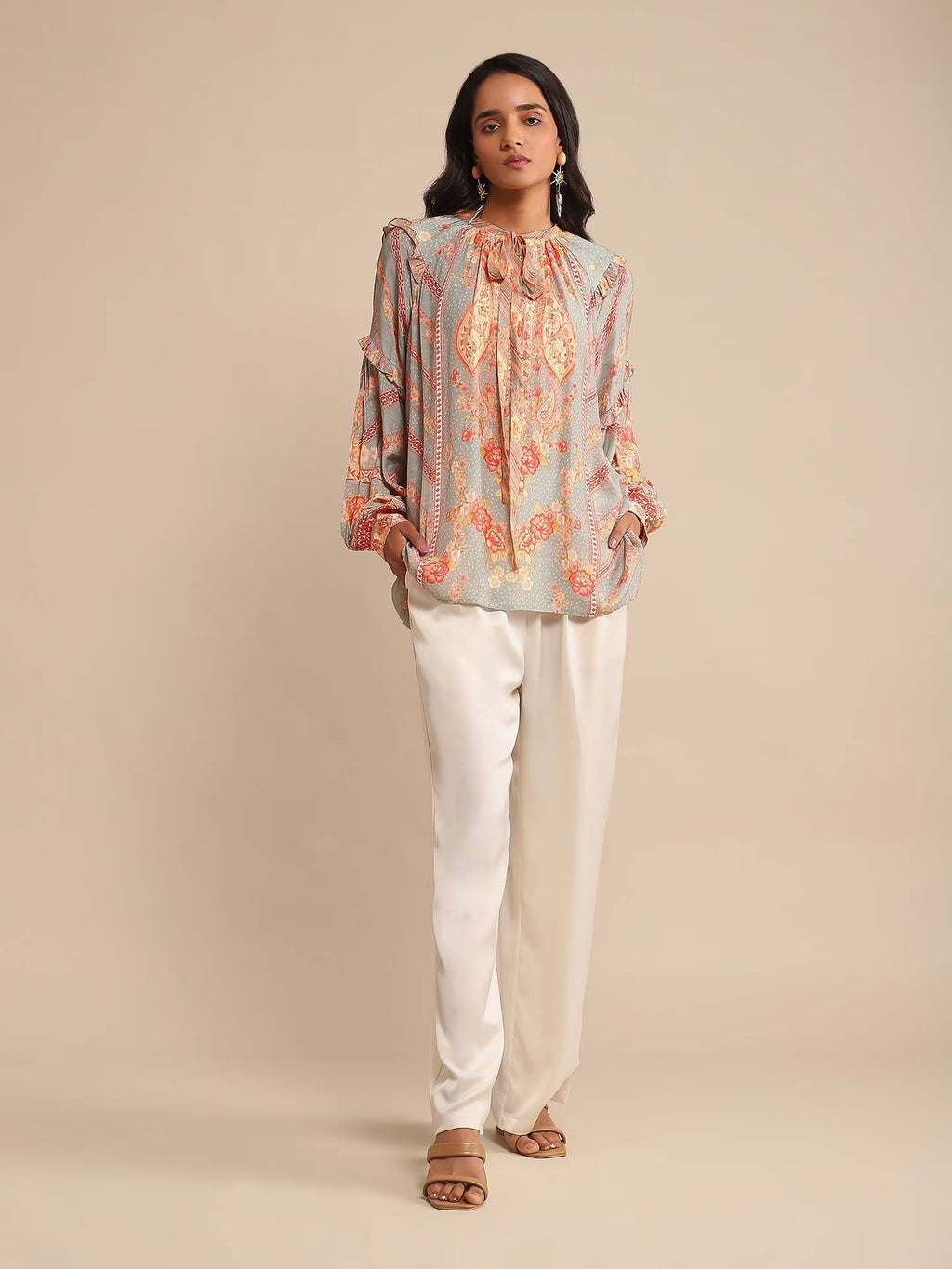 Woman in a multicolor printed high-neck kurta with full sleeves, paired with white straight pants and sandals, standing against a beige background