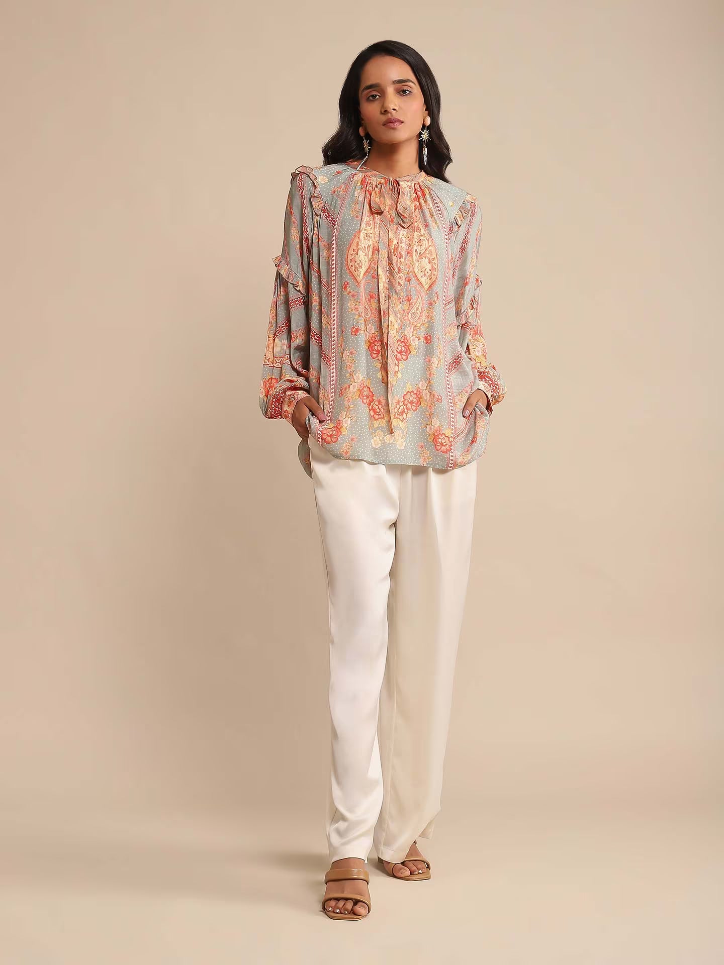 Woman in a multicolor printed high-neck kurta with full sleeves, paired with white straight pants and sandals, standing against a beige background