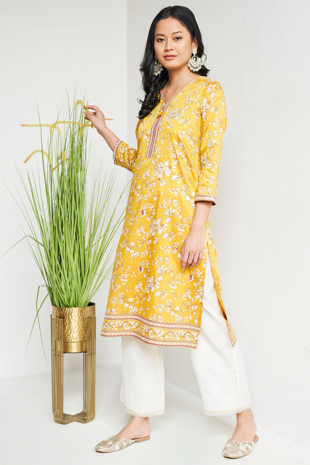 Woman wearing a yellow floral kurta with white pants standing next to a plant on a white background