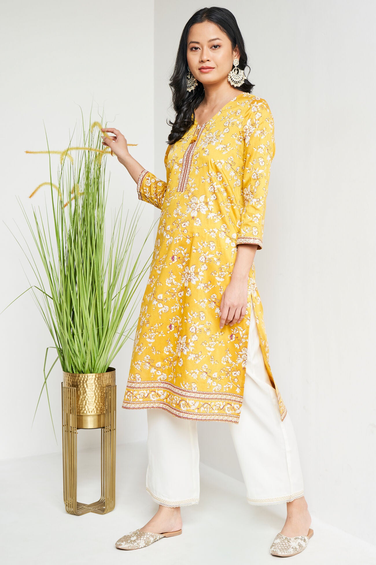 Woman wearing a yellow floral kurta with white pants standing next to a plant on a white background