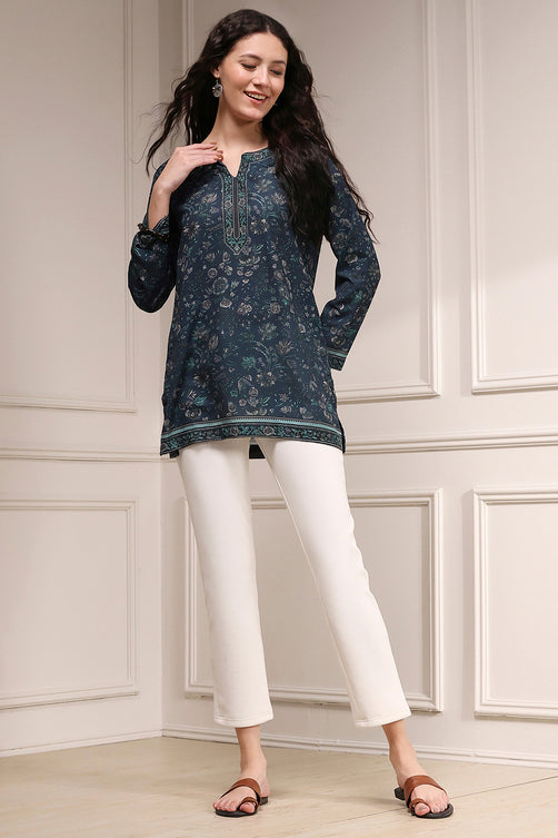 Woman wearing a dark blue floral top and white pants against a light paneled wall.