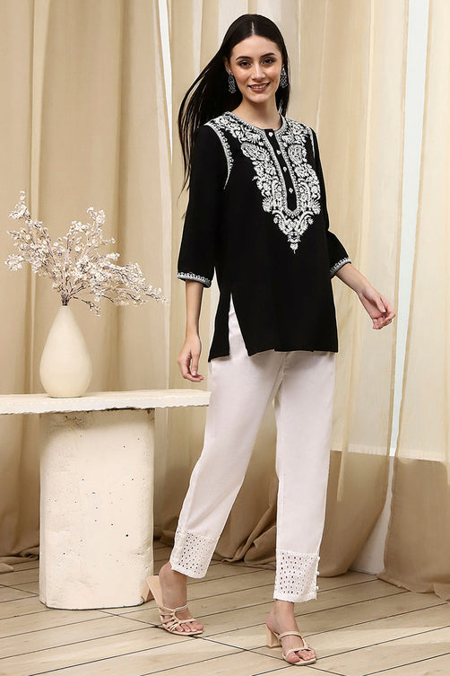 Woman wearing a black embroidered top and white pants standing in a room with beige curtains and a vase with flowers.