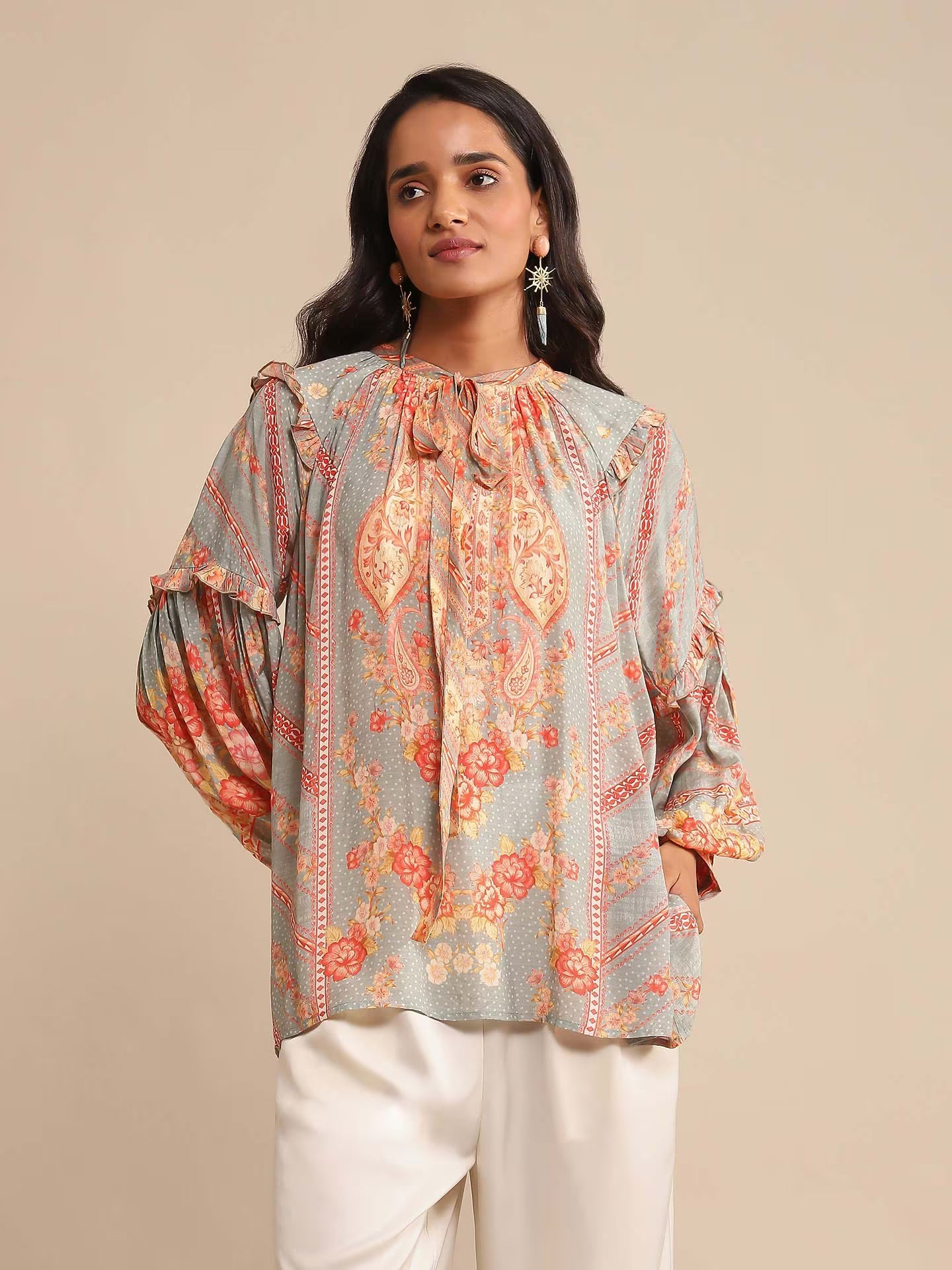 Woman in a multicolor printed high-neck kurta with full sleeves, paired with white straight pants and sandals, standing against a beige background