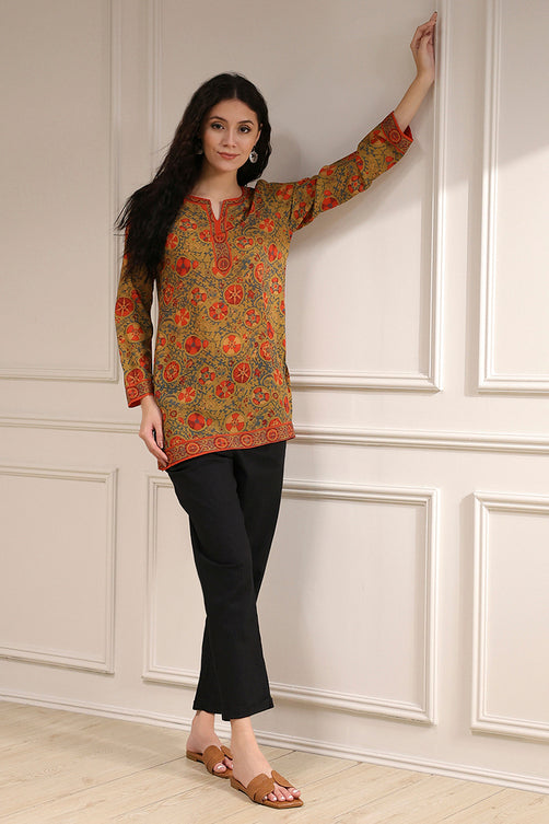 Woman wearing a patterned top and black pants against a white paneled wall.