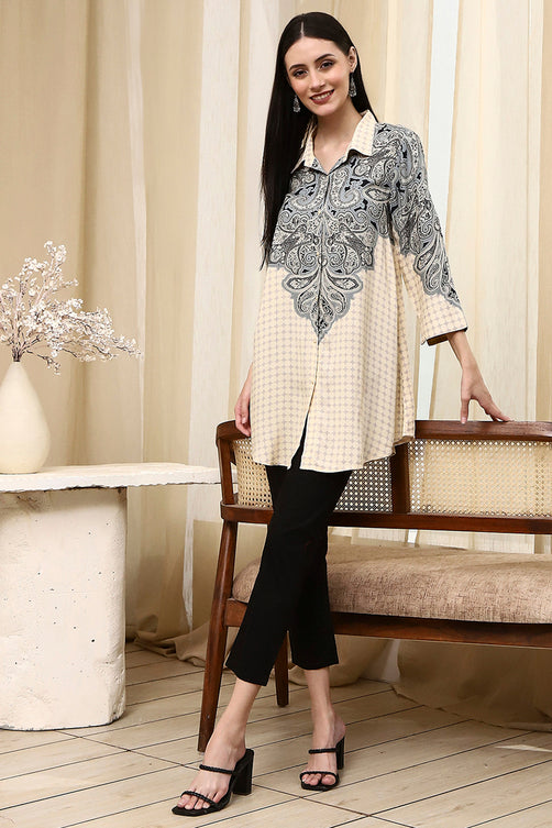 Woman in a stylish outfit with a patterned top and black pants, standing in a room with a wooden chair and decorative elements.