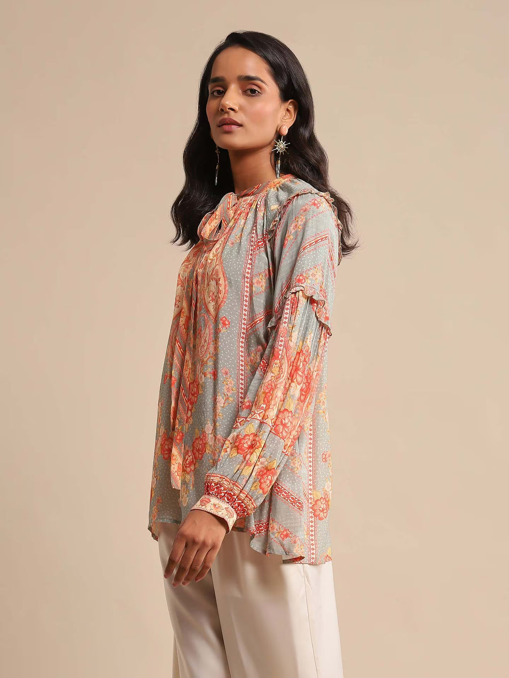 Woman in a multicolor printed high-neck kurta with full sleeves, paired with white straight pants and sandals, standing against a beige background