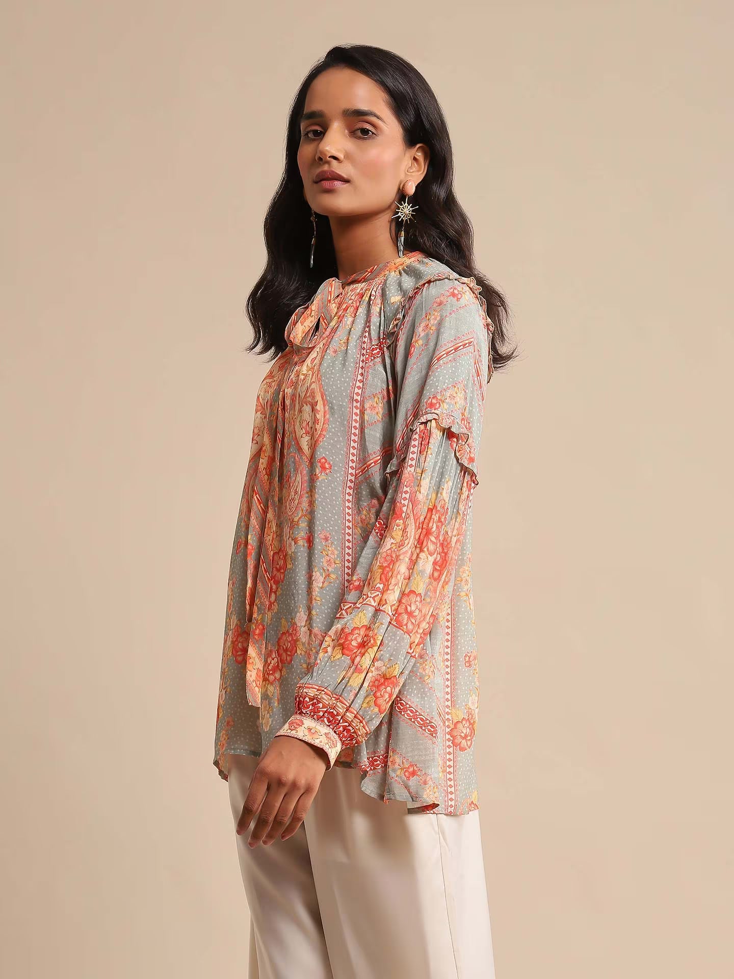 Woman in a multicolor printed high-neck kurta with full sleeves, paired with white straight pants and sandals, standing against a beige background