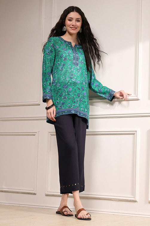 Woman wearing a green floral top and black pants against a white paneled wall.