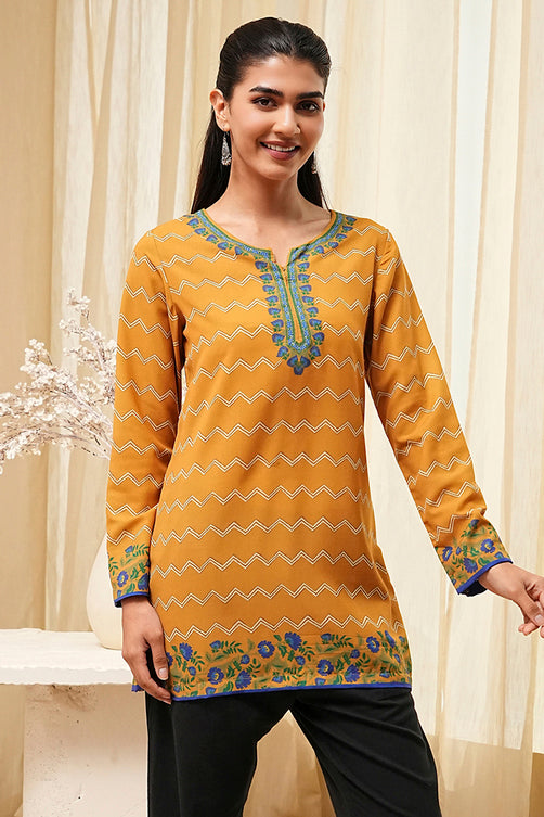 Woman wearing a yellow patterned top with black pants against a beige curtain background