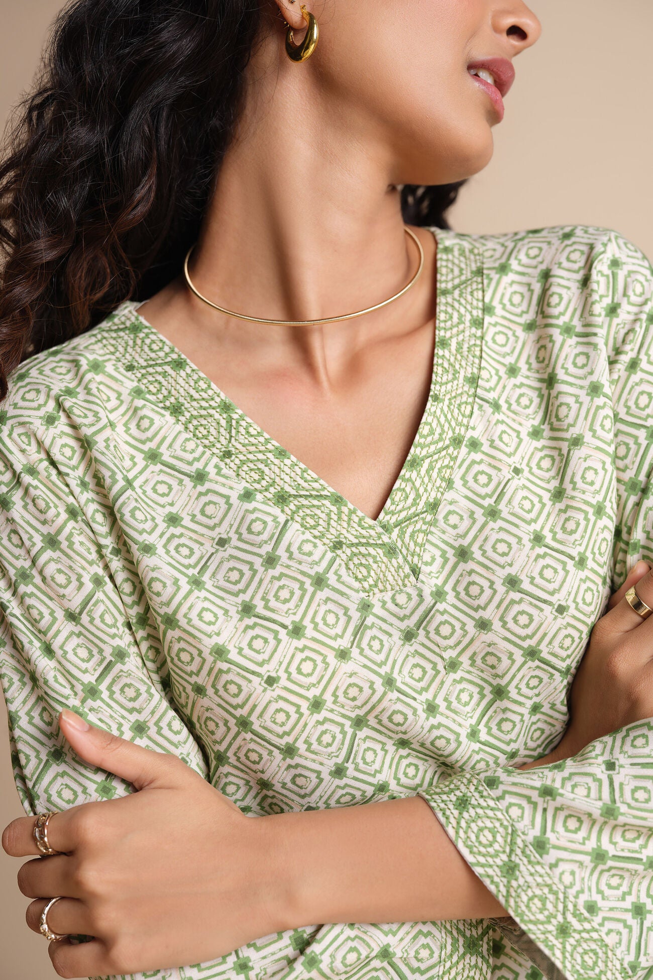 Woman wearing a green patterned top with gold jewelry on a beige background