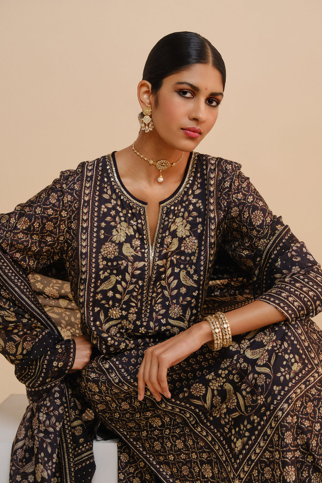 Woman wearing a black and gold embroidered traditional outfit with jewelry on a beige background