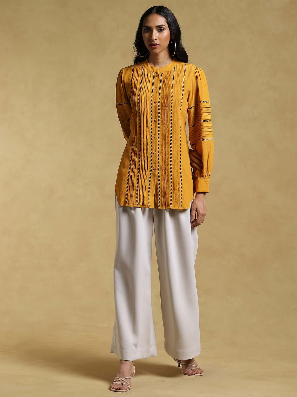 Woman wearing a mustard yellow top and white pants against a beige background