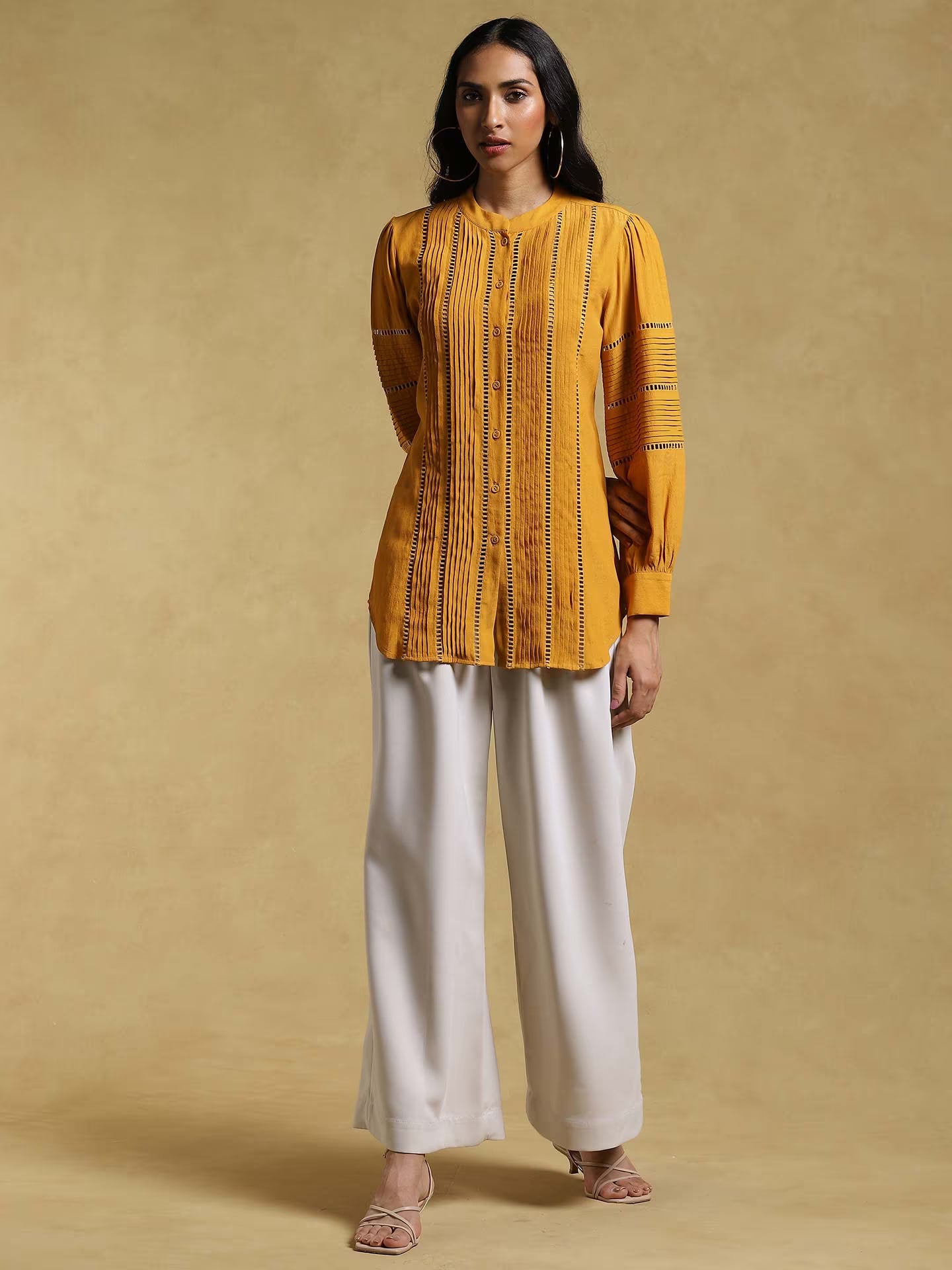Woman wearing a mustard yellow top and white pants against a beige background